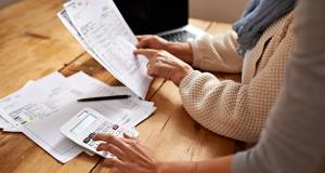 Stock photo of two people reviewing medical bills at a table. On the table are a laptop, a calculator, and several papers. 